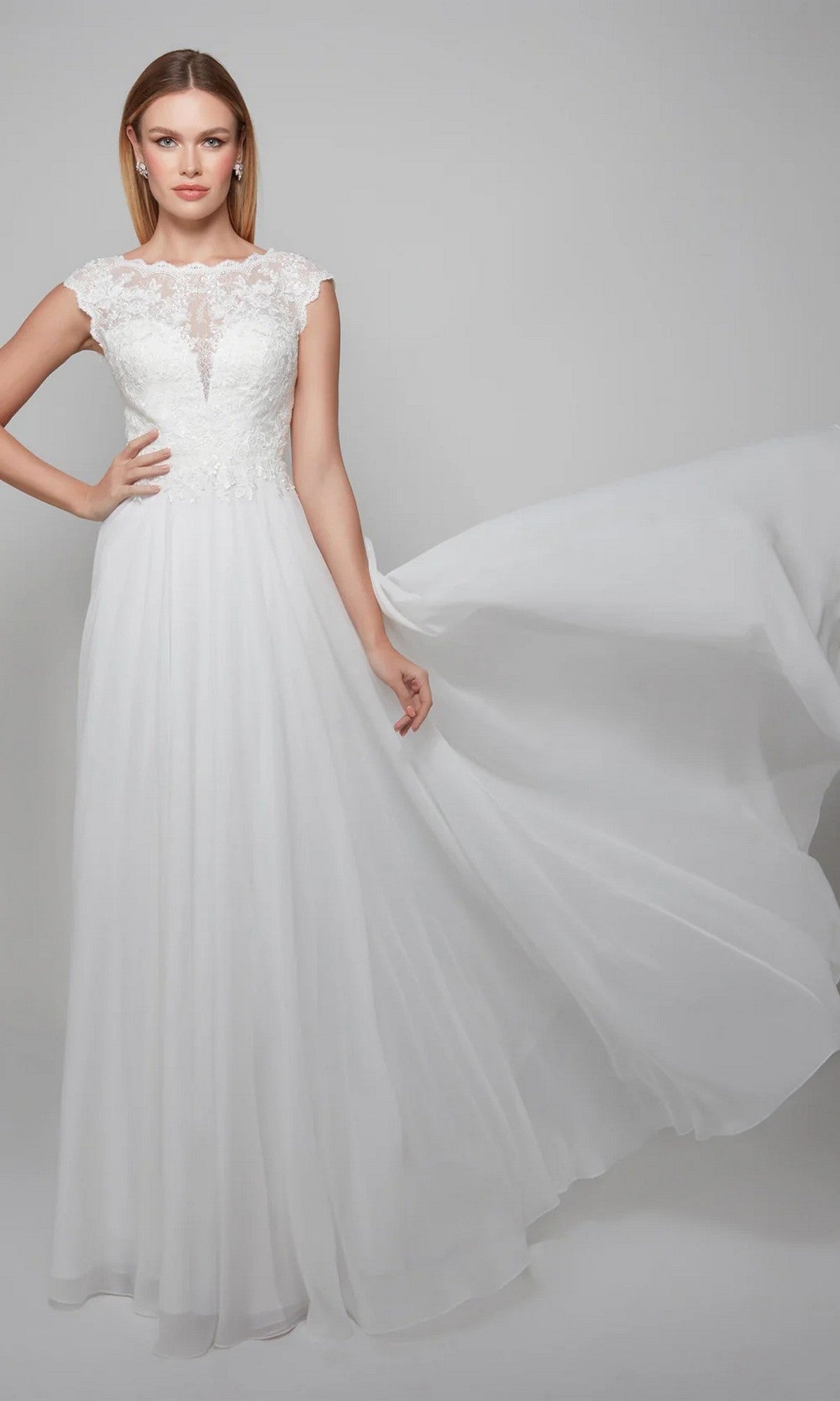 A woman models the Long White Dress: Alyce 7073 in Diamond White, featuring lace embroidery and a chiffon A-line skirt, as she holds out one side of the flowing skirt against a plain light background.